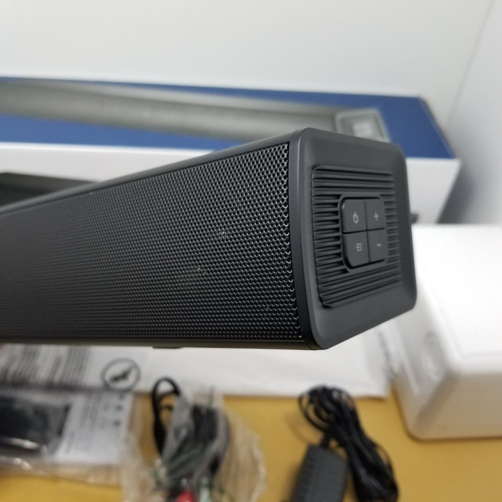 Black speaker bar with control buttons on the side, white surface with packaging in the background