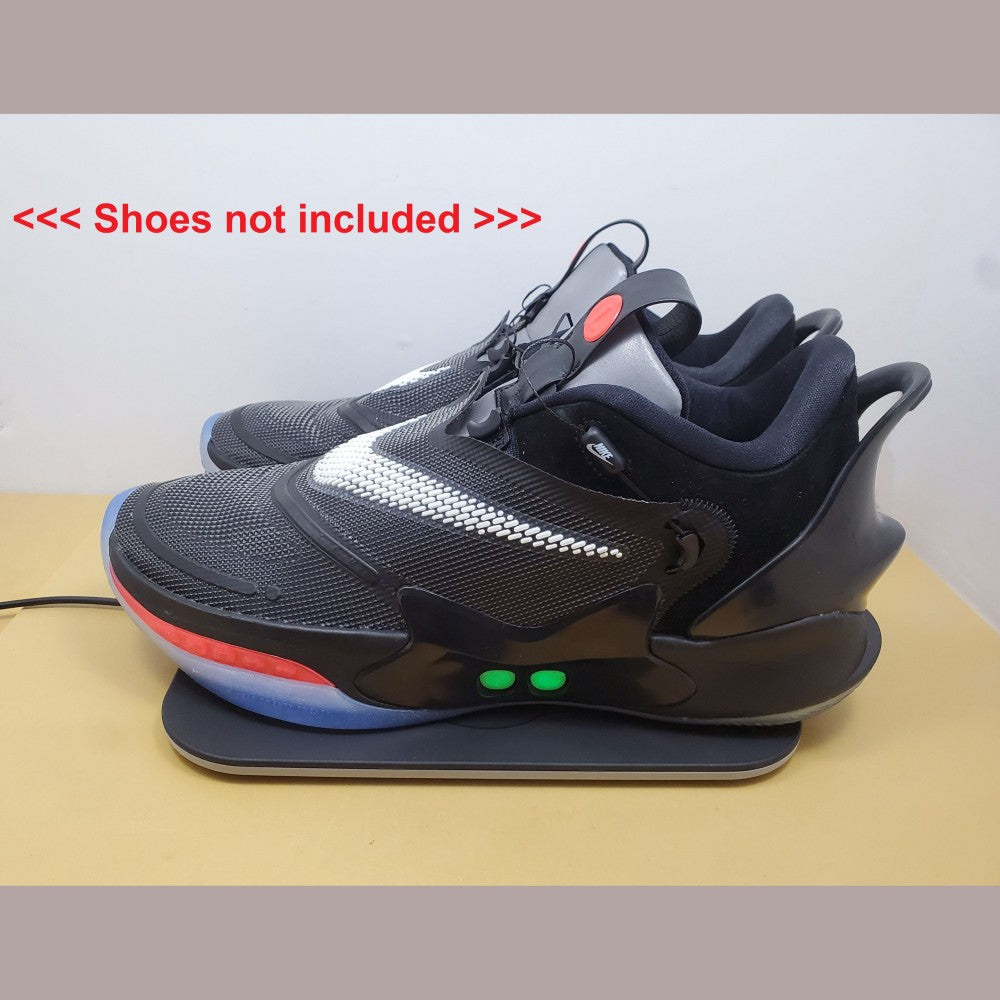 Black Nike Adapt shoes with two green lights on the side, sitting on Charging Mat on a beige surface. Also notation saying "shoes not included" can be seen next to shoes.