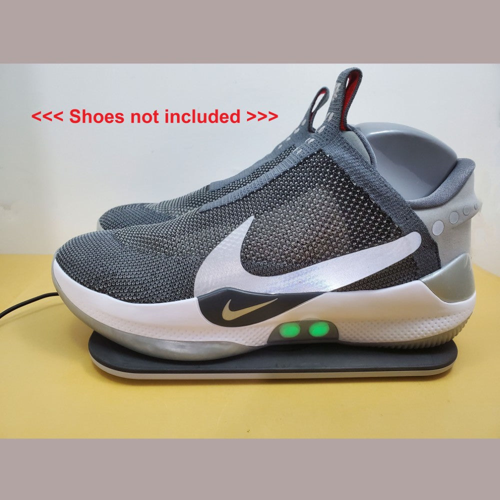Gray Nike Adapt shoes with two green lights on the side, sitting on Charging Mat on a beige surface. Also notation saying "shoes not included" can be seen next to shoes.