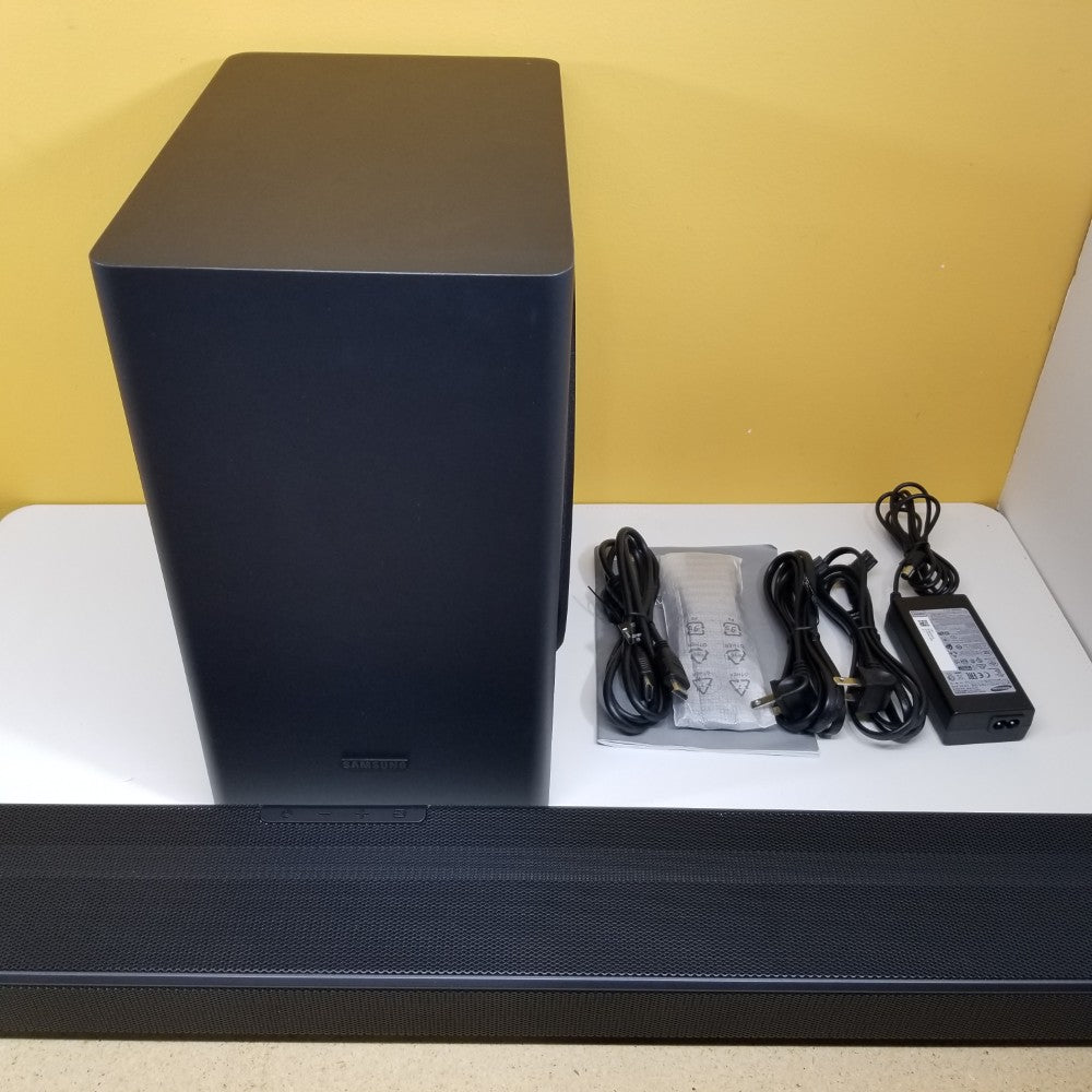 Samsung HW-Q60T subwoofer & soundbar with cables and power adapter on a white surface against a yellow wall.